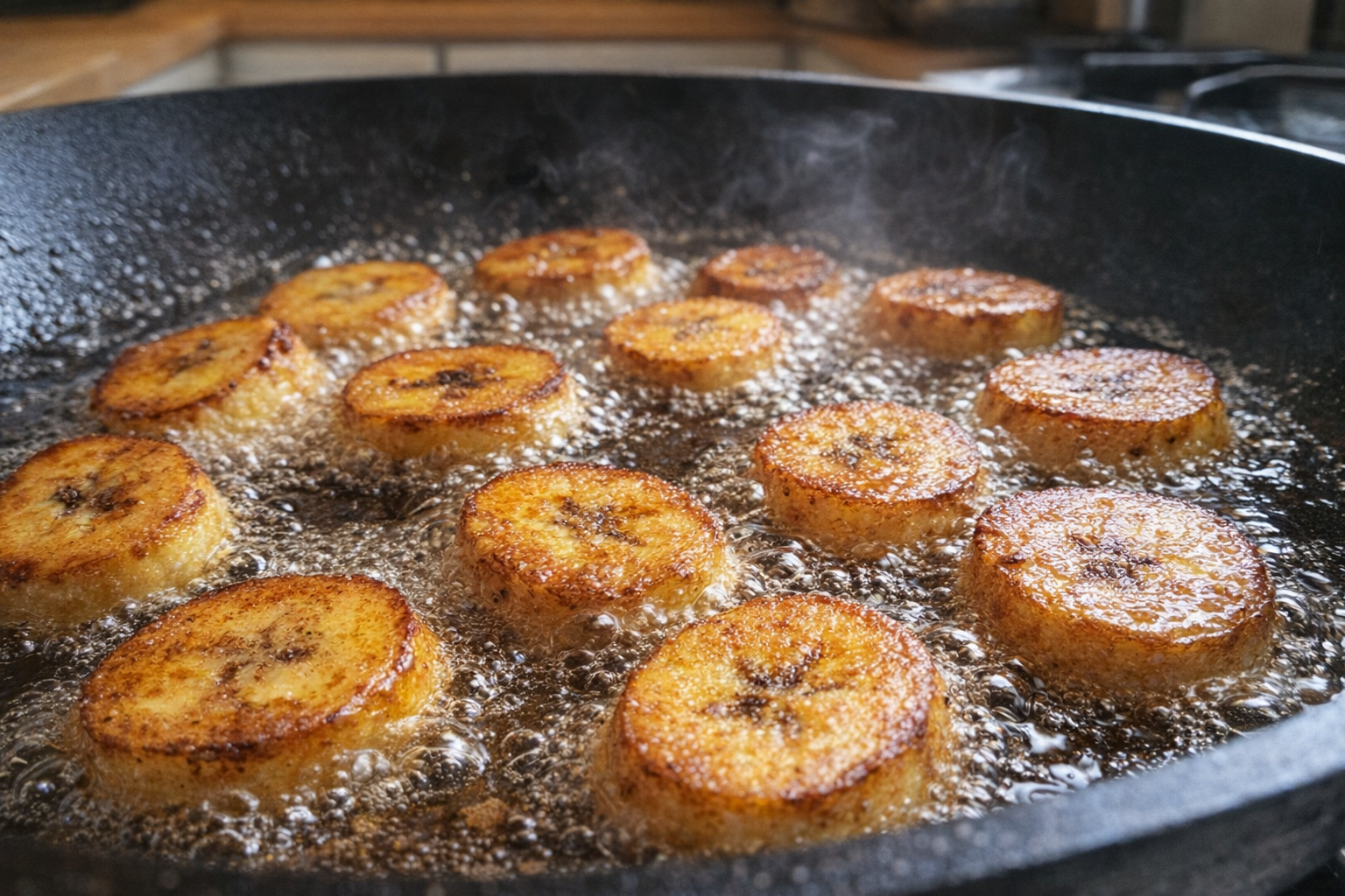 An action shot of plantain slices sizzling in a hot skillet, with oil bubbling around the edges and the plantains turning golden brown